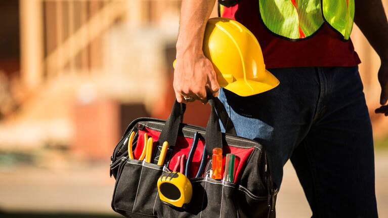 A construction worker busy working at a job site. He holds a tool box full of tools and a hard hat. Framed house, building in background. He is wearing a safety vest.