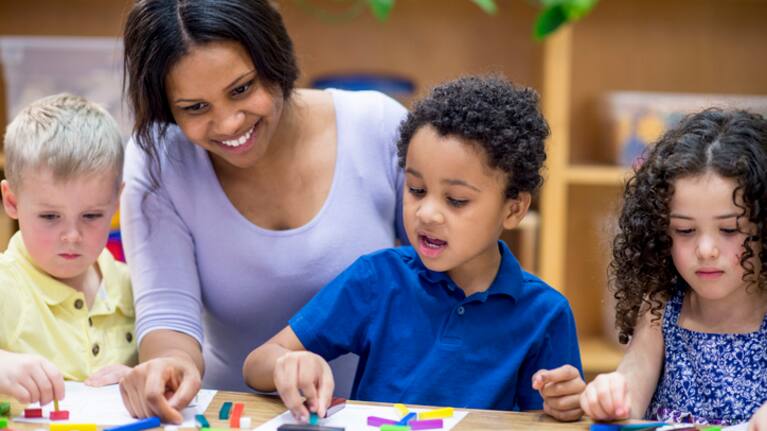 A multi-ethnic group of young children are indoors in a school. They are wearing casual clothing. They are sitting at a table and playing with blocks along with their teacher.